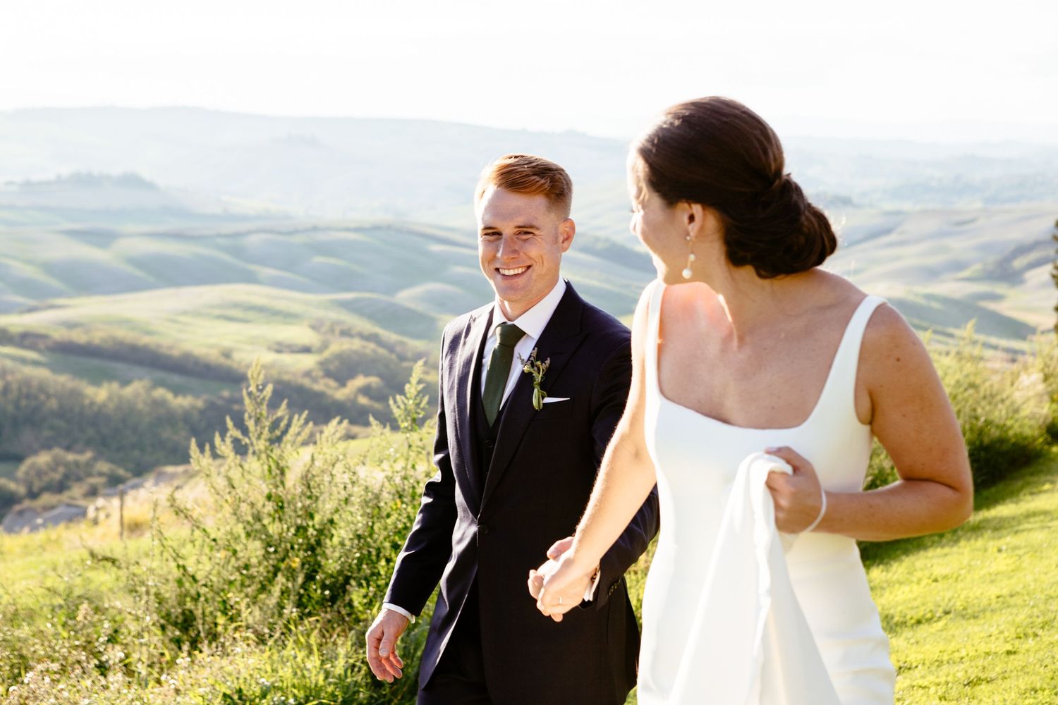 The bride and groom walk together around the grounds of the Lazy Olive with the rolling hills of Siena in the background