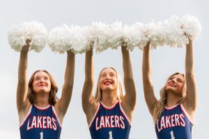 Three high school dance team dancers in matching Lakers uniforms hold white pom poms overhead while smiling jubilantly at the camera.
