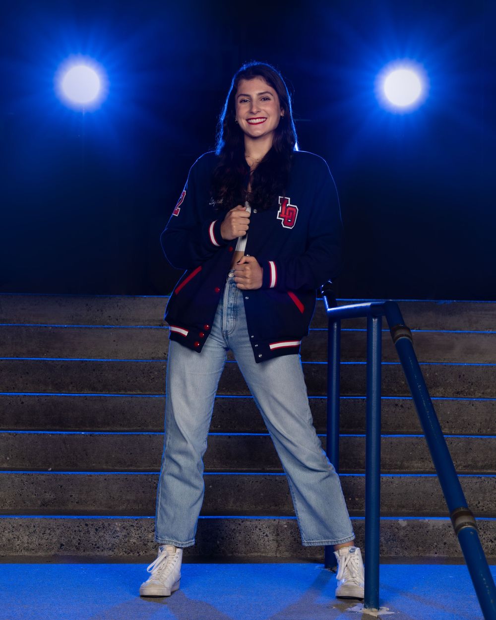 High School Dancer wearing a varsity letterman jacket and jeans poses on steps with blue spotlights in the background.
