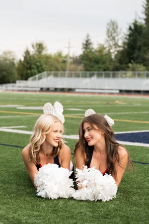 Two High School Dancers lying on football field with white pom poms and smiling during outdoor photoshoot.