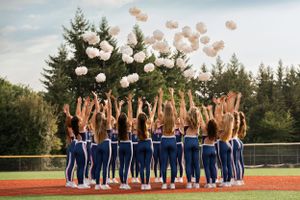 High School dance team dancers in blue uniforms throws pom poms into the air on a sports field surrounded by evergreen trees.