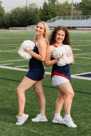 Two cheerleaders in navy uniforms pose with white pom-poms on a football field at a stadium.