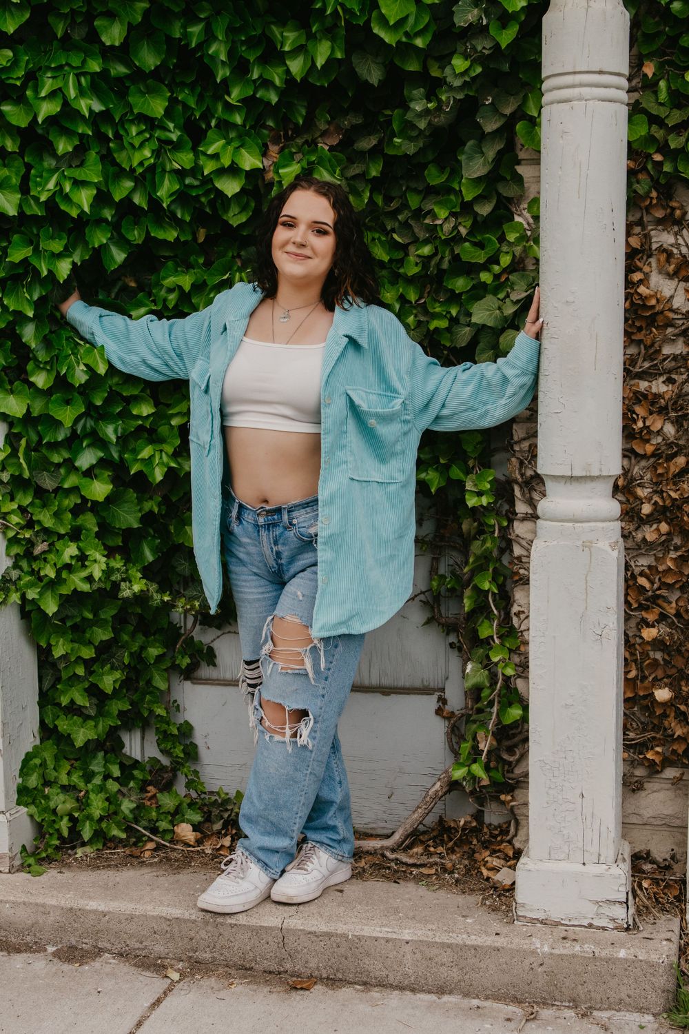 Young woman in teal jacket and ripped jeans posing by ivy wall.