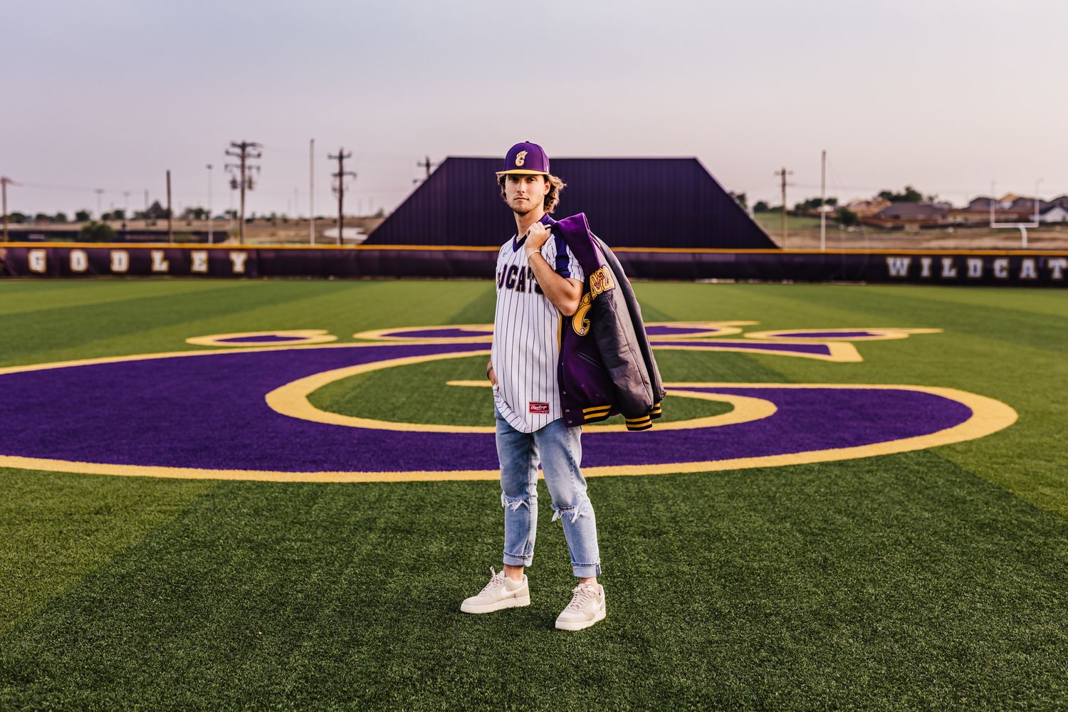 A baseball player stands on a purple and gold field with sports equipment.