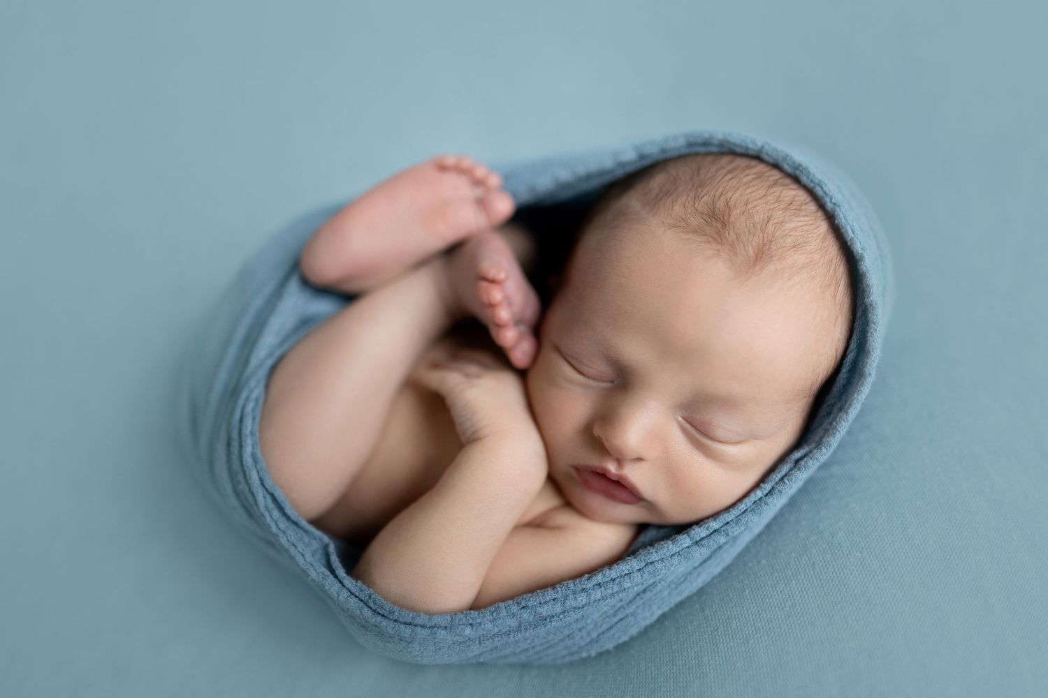 Peaceful sleeping infant curled up in soft blue wrap against blue background.