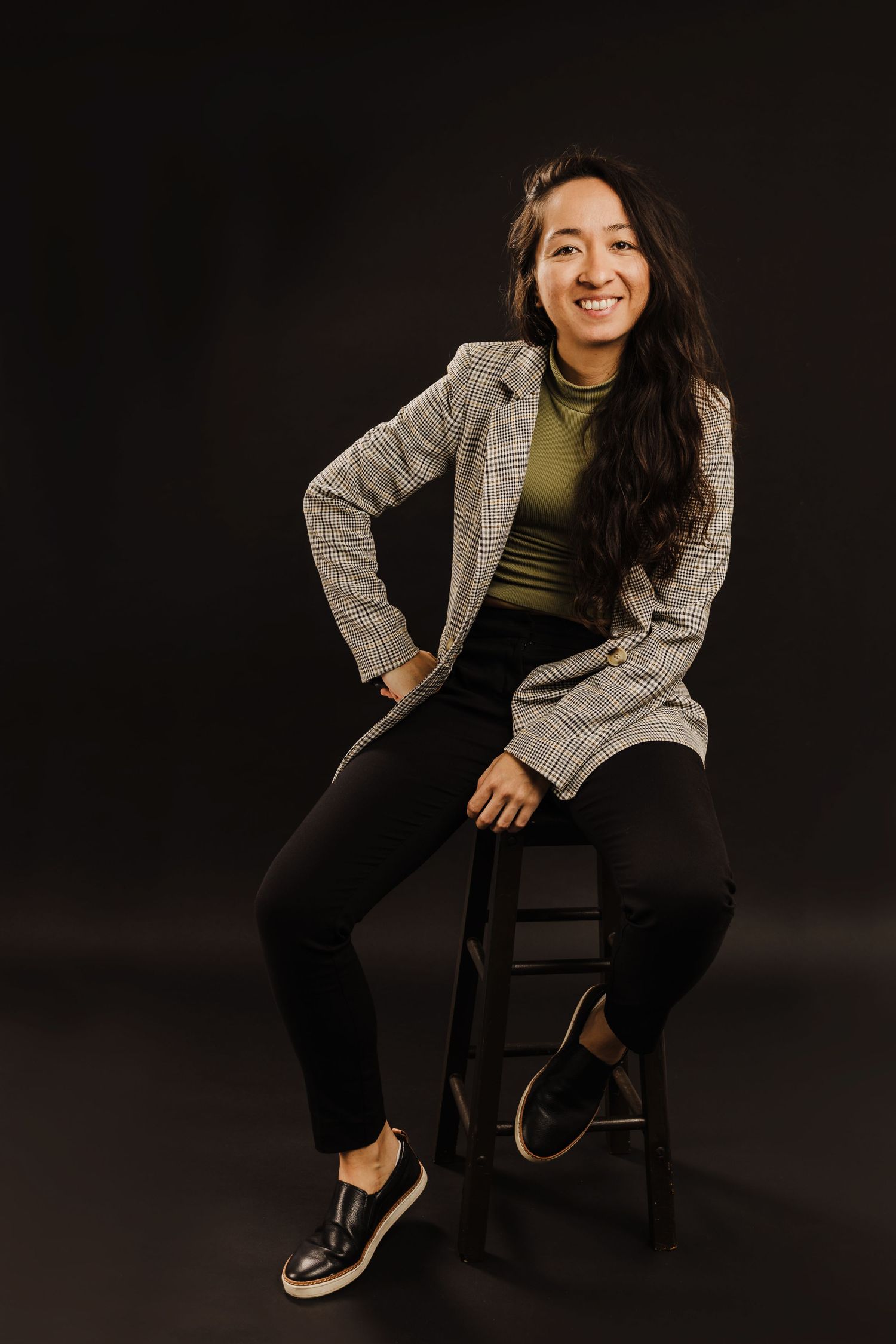 Woman in business casual attire sitting on stool against dark background wearing plaid blazer.