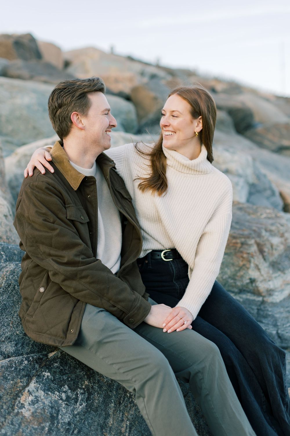 A romantic couple laughs together while sitting on coastal rocks during a sunset photoshoot in Montauk, Long Island, New York.