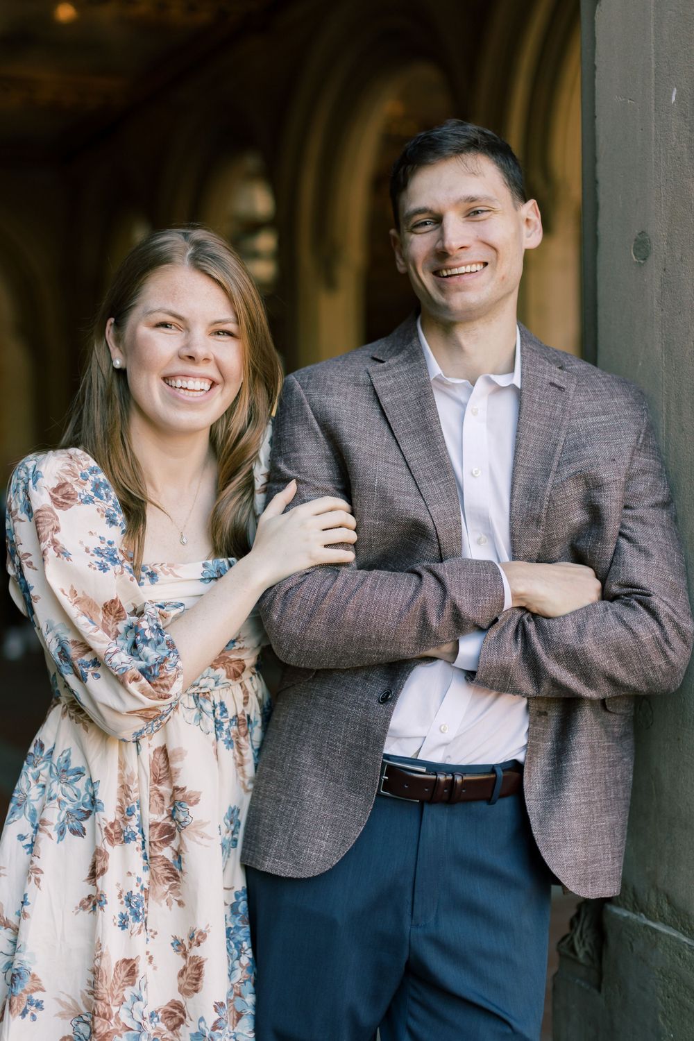 A couple in casual formal wear poses smiling against a stone archway with a floral dress and gray blazer.