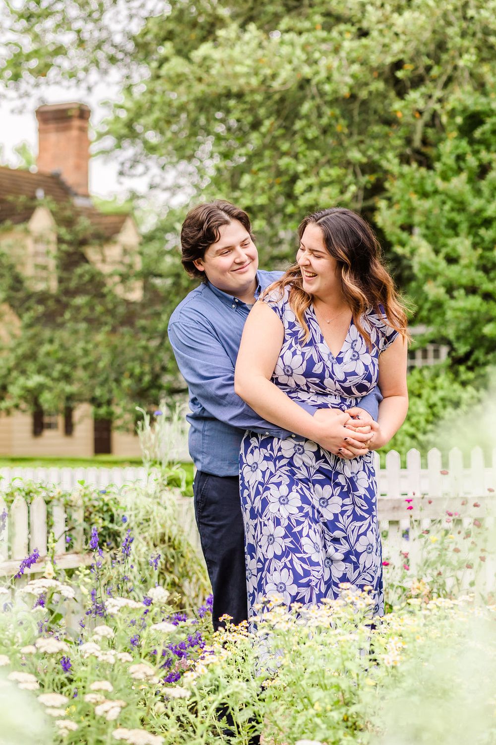 A couple embraces in a flowering garden with white picket fence and brick house in background.