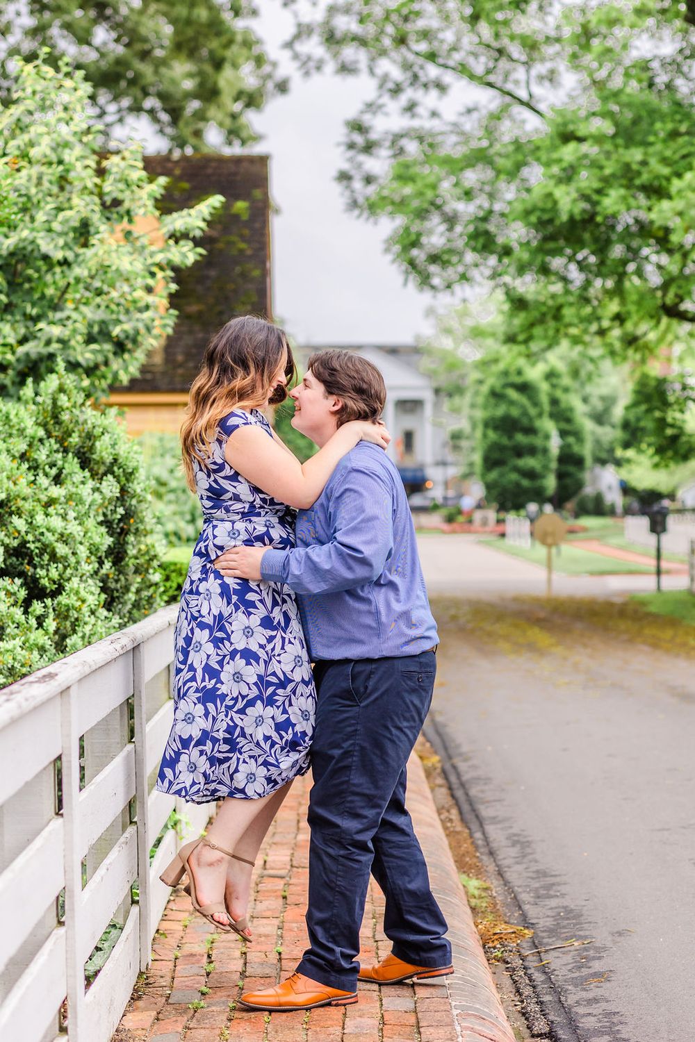 A couple embraces on a brick pathway with white fencing and greenery.