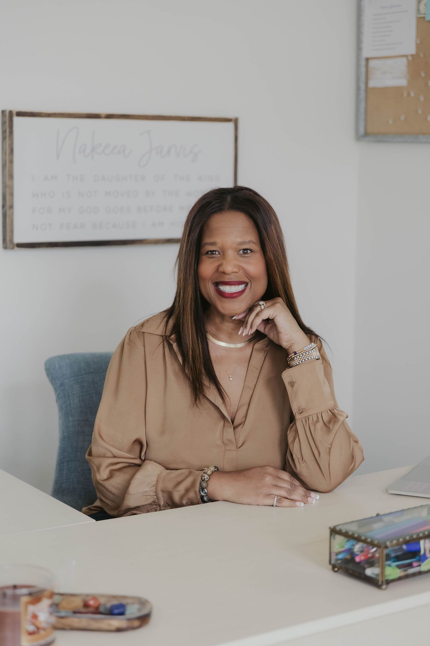 Business professional in a tan blouse smiles joyfully while seated at a desk in a bright office space.