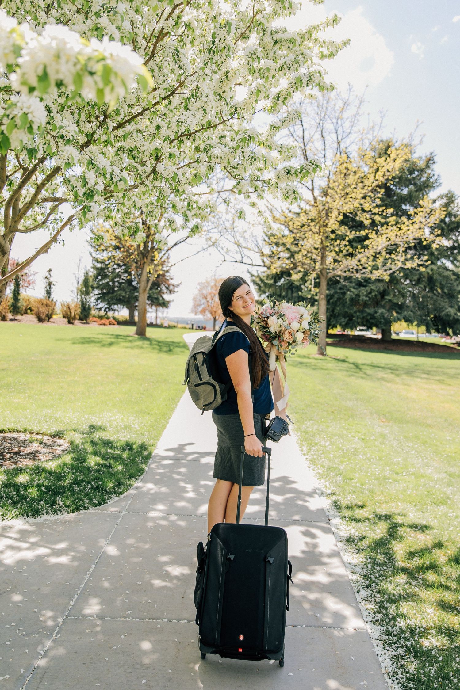 Person with luggage and backpack standing on sunny path under blooming spring trees.