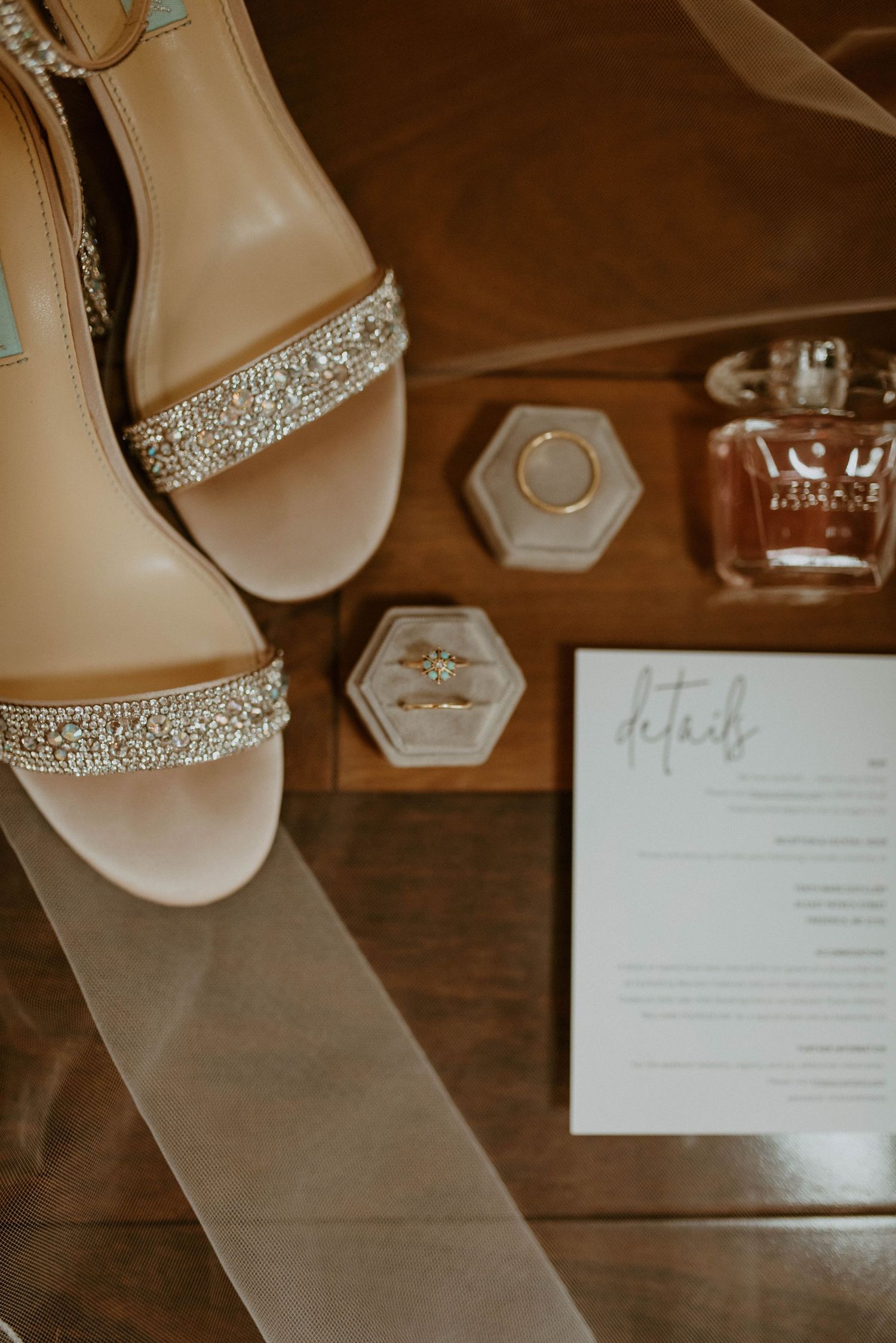 Wedding details flatlay with sparkly heels, perfume bottle, and invitation on wooden surface.