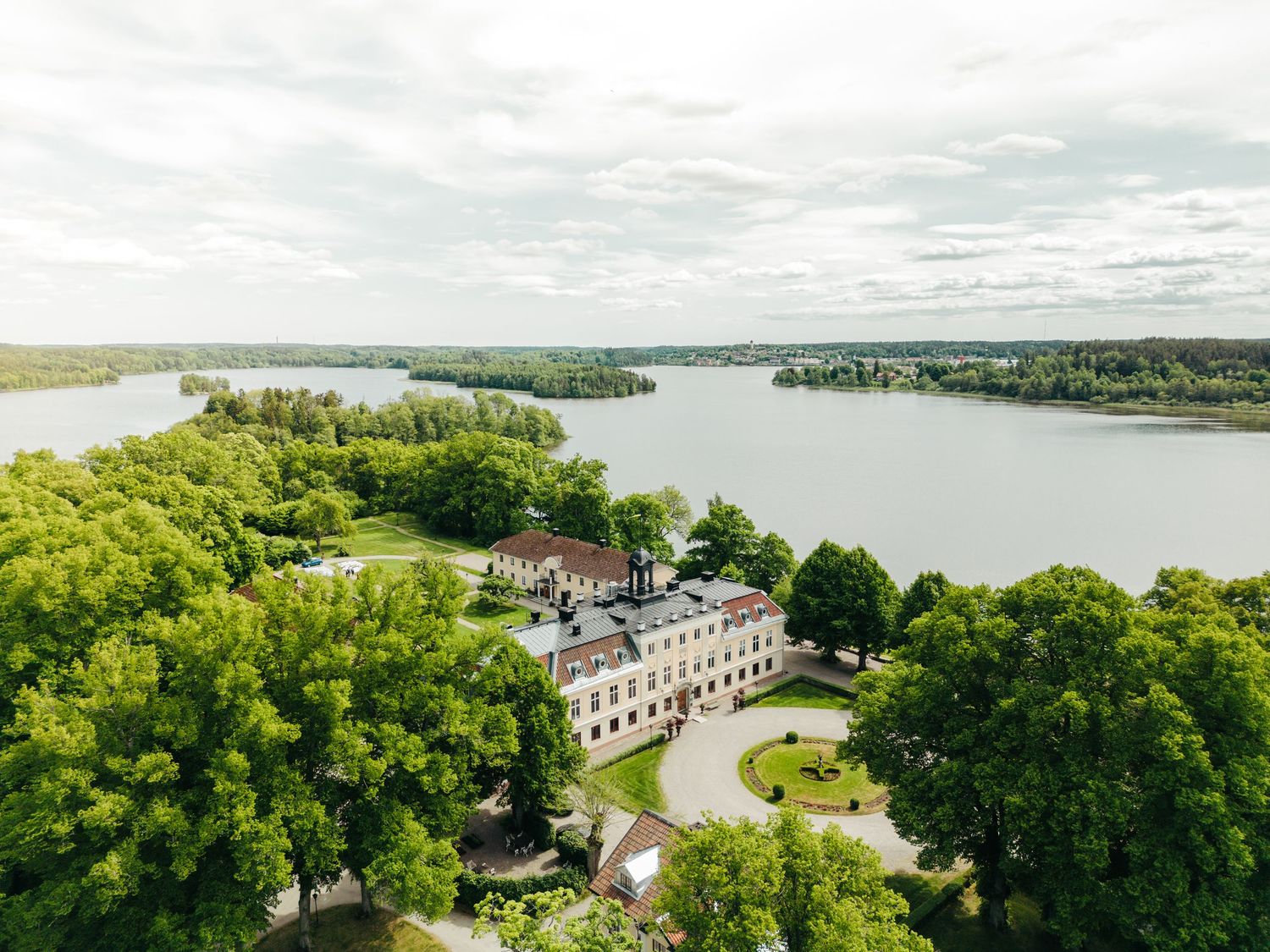 Aerial view of a grand mansion surrounded by lush trees and a winding lake on a cloudy day.