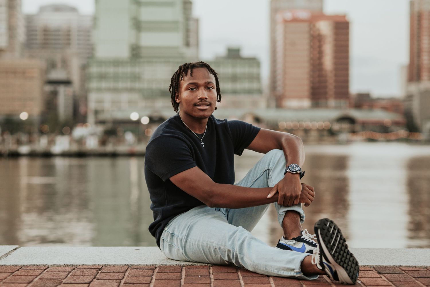 Person in casual wear sits by waterfront with city skyline in background.