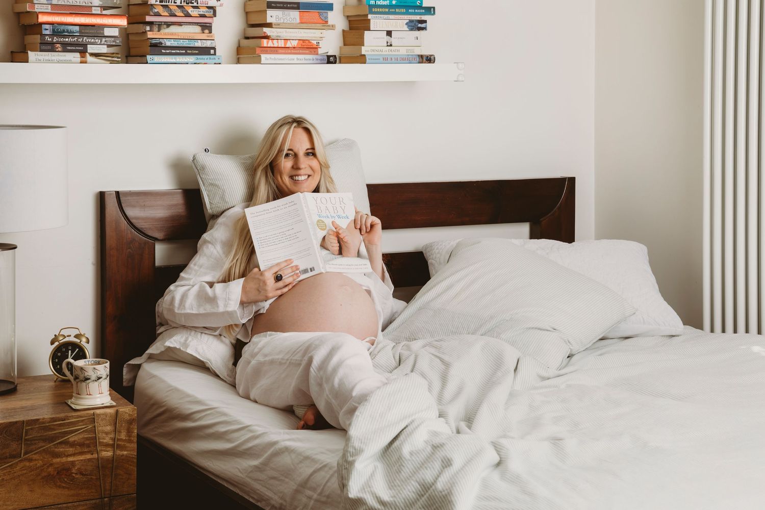 Pregnant woman reading in bed surrounded by white linens and books on shelf.