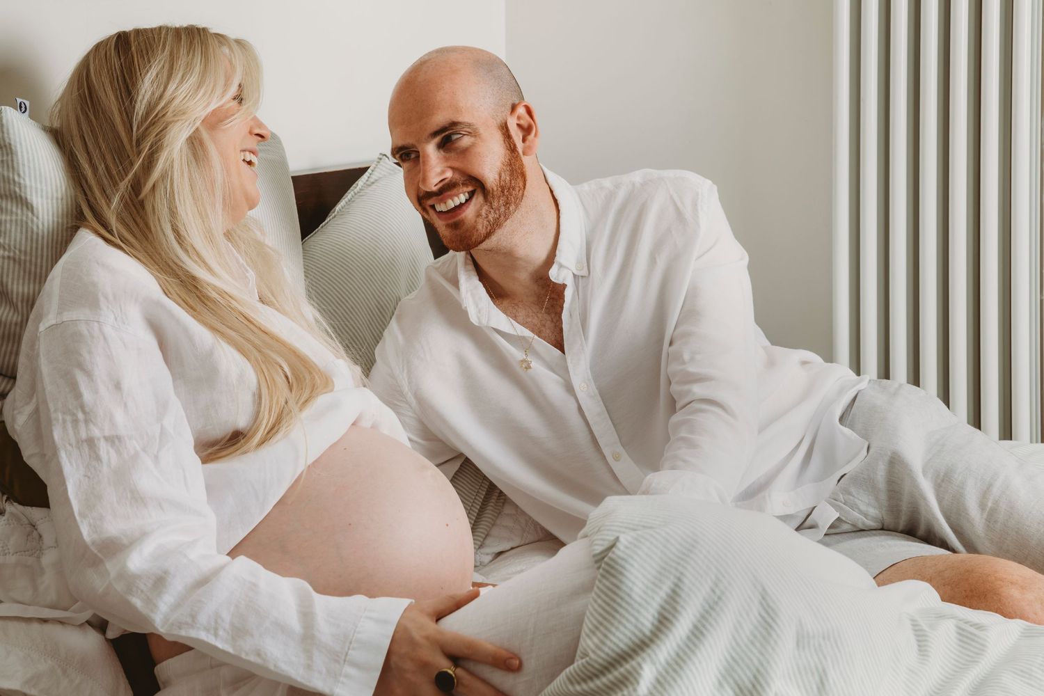 Intimate maternity photo of couple in white clothing relaxing on bed together.