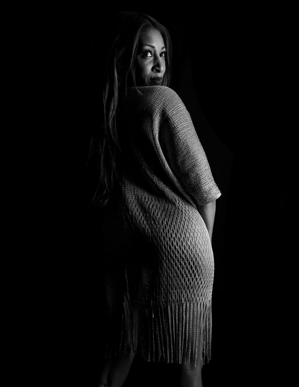 Dramatic black and white artistic portrait showing a figure in a knit sweater dress against a dark background.