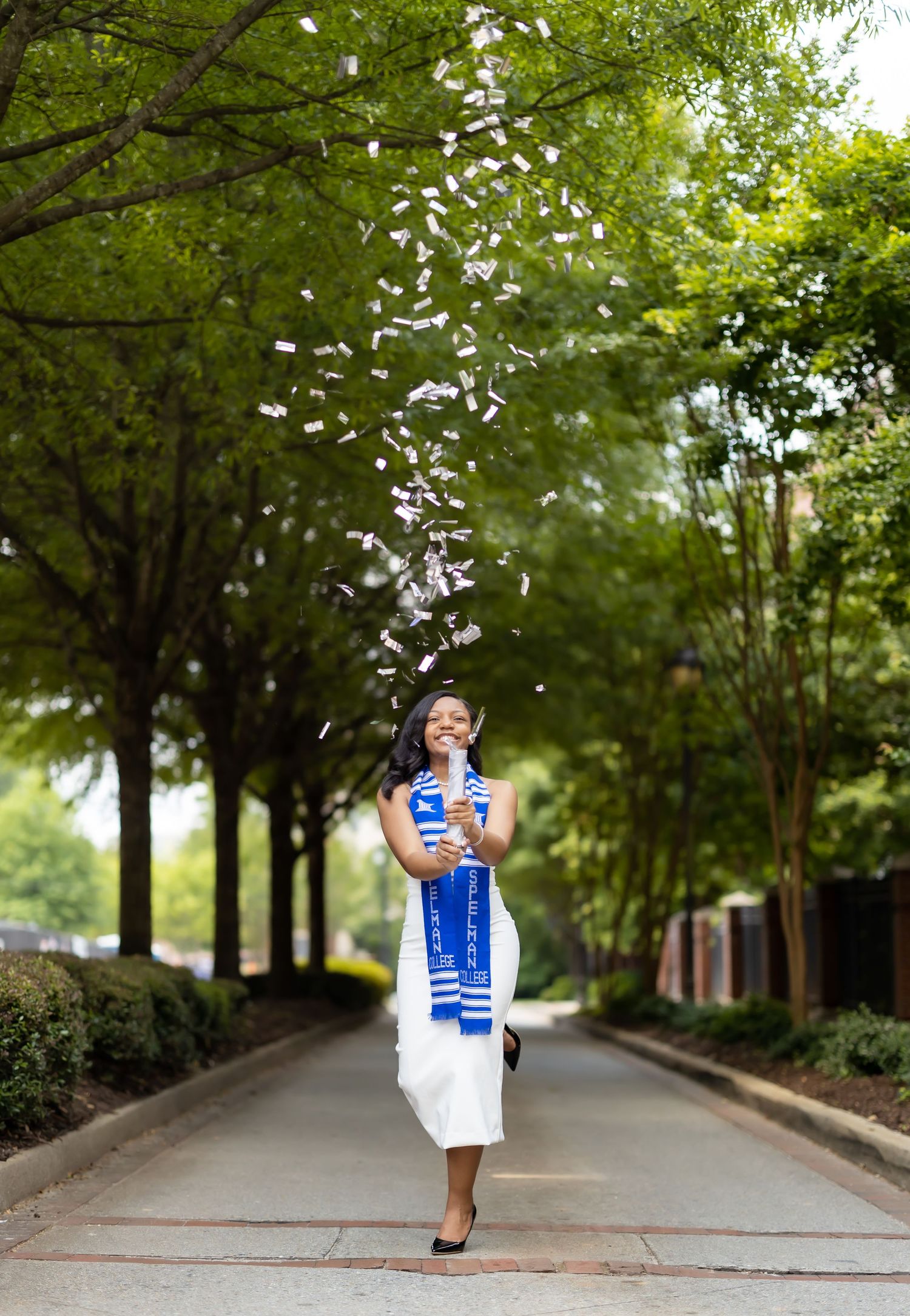 A person in blue and white attire throws flower petals while walking down a tree-lined path.