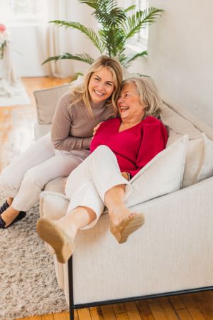 A woman and her elderly mother share joyful laughter while relaxing together on a comfortable couch with a palm plant in the background.