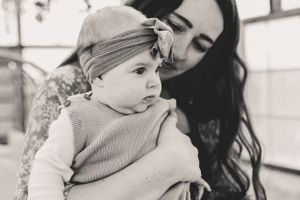 A black and white photograph shows a baby wearing a knit sweater and headband being held lovingly against a bright background.