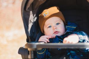 A baby wearing a brown winter hat sits in a stroller with a blurred background in autumn sunlight.