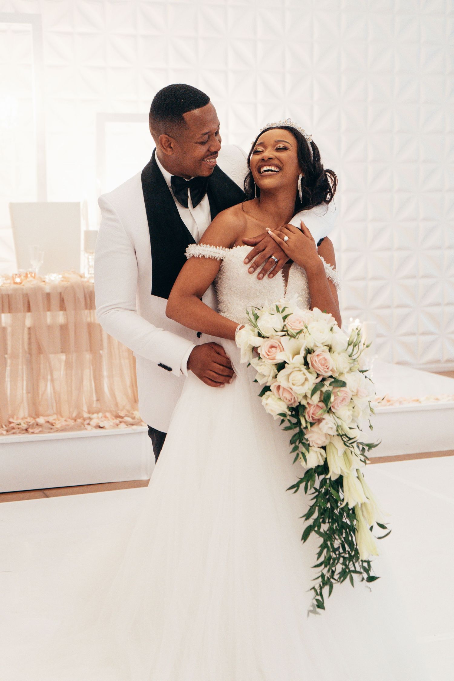 A joyful bride holds a cascading bouquet as her groom embraces her in an elegant white venue.