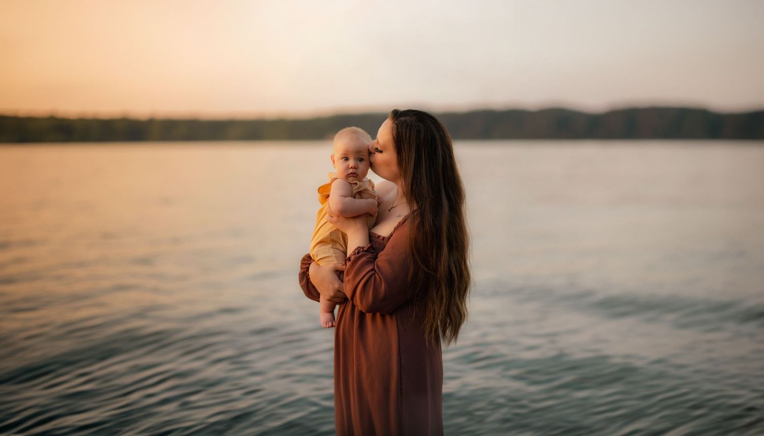 A figure in a rust-colored dress holds a small dog while standing by a tranquil lake at sunset.
