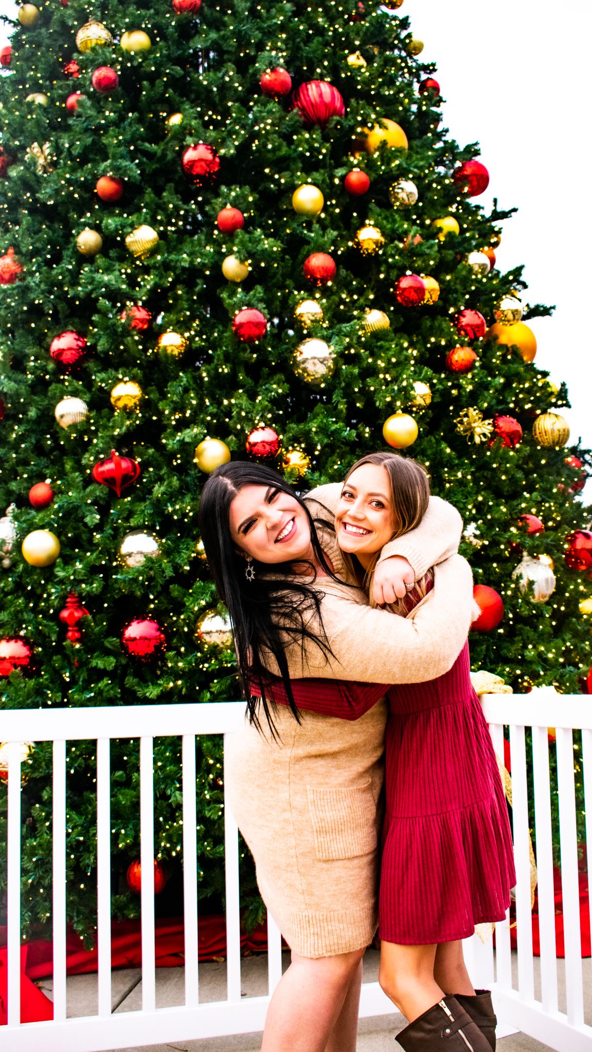 Two people posing in front of large decorated Christmas tree with red and gold ornaments.