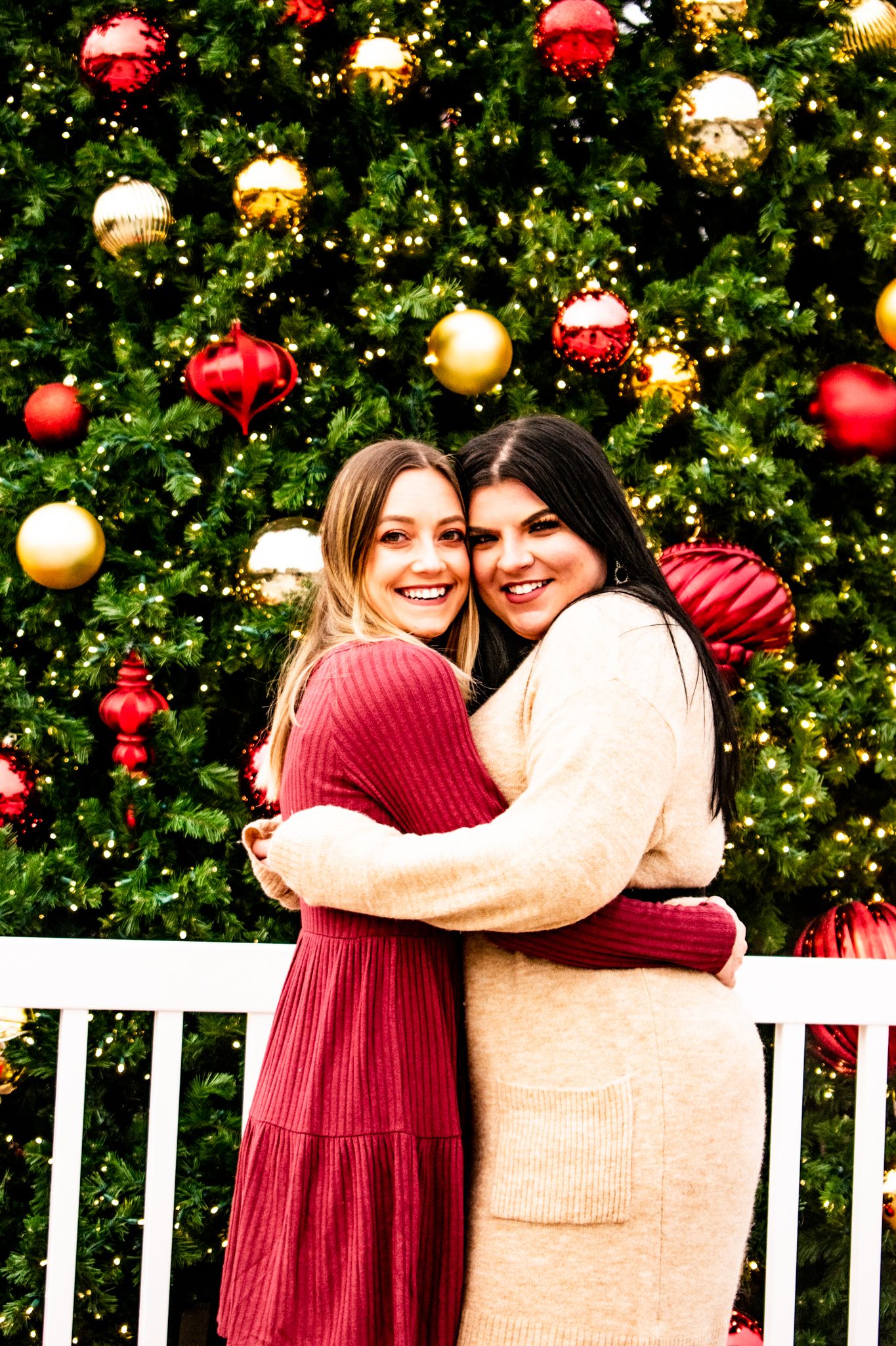 Friends embracing in front of festive Christmas tree with red and gold decorations.