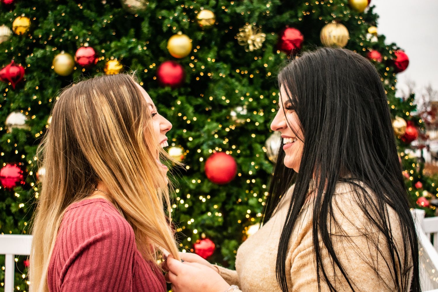 Two friends share laughter in front of decorated Christmas tree with twinkling lights.