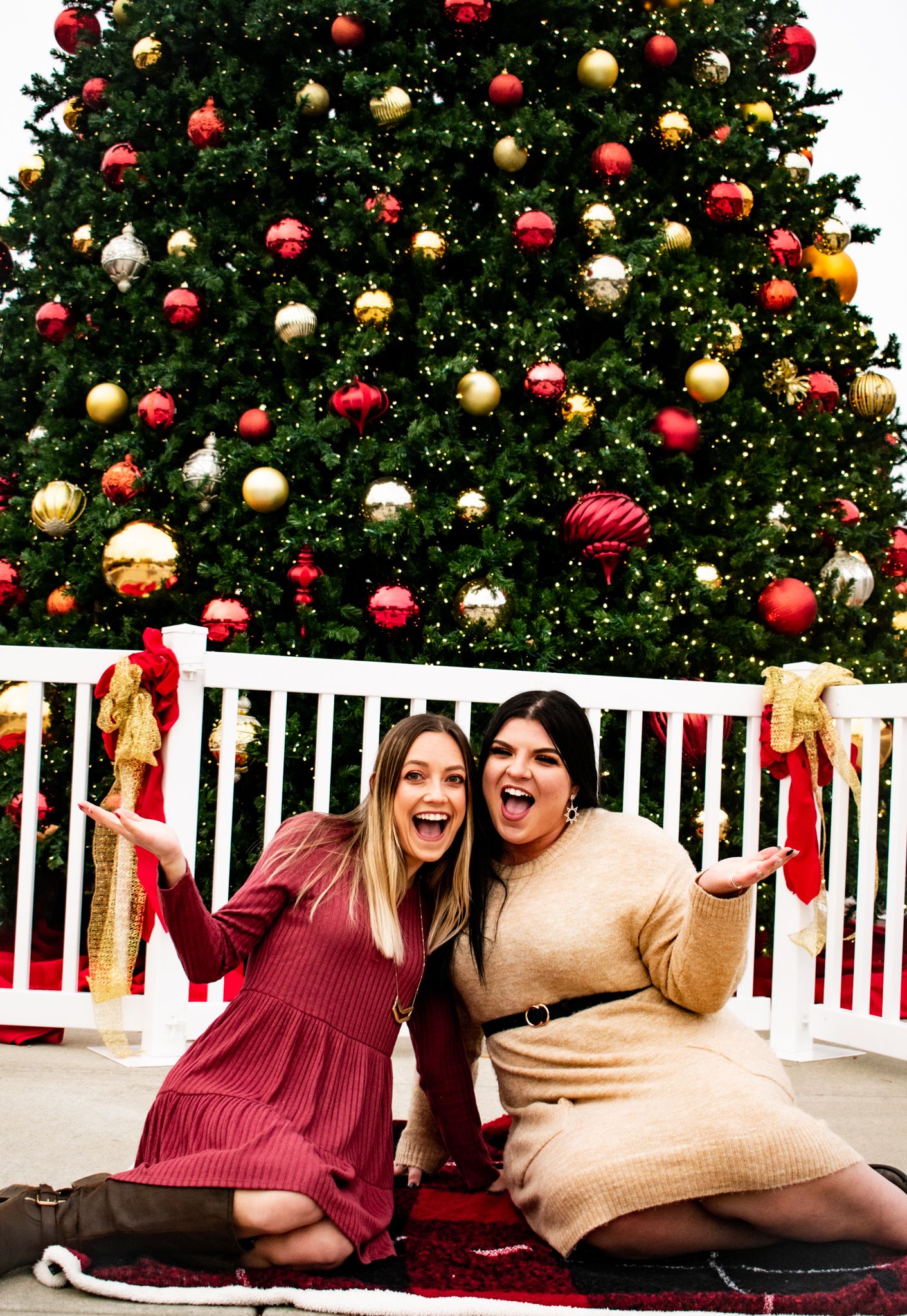Two friends share joyful laughter while sitting on a red blanket in front of a decorated Christmas tree.