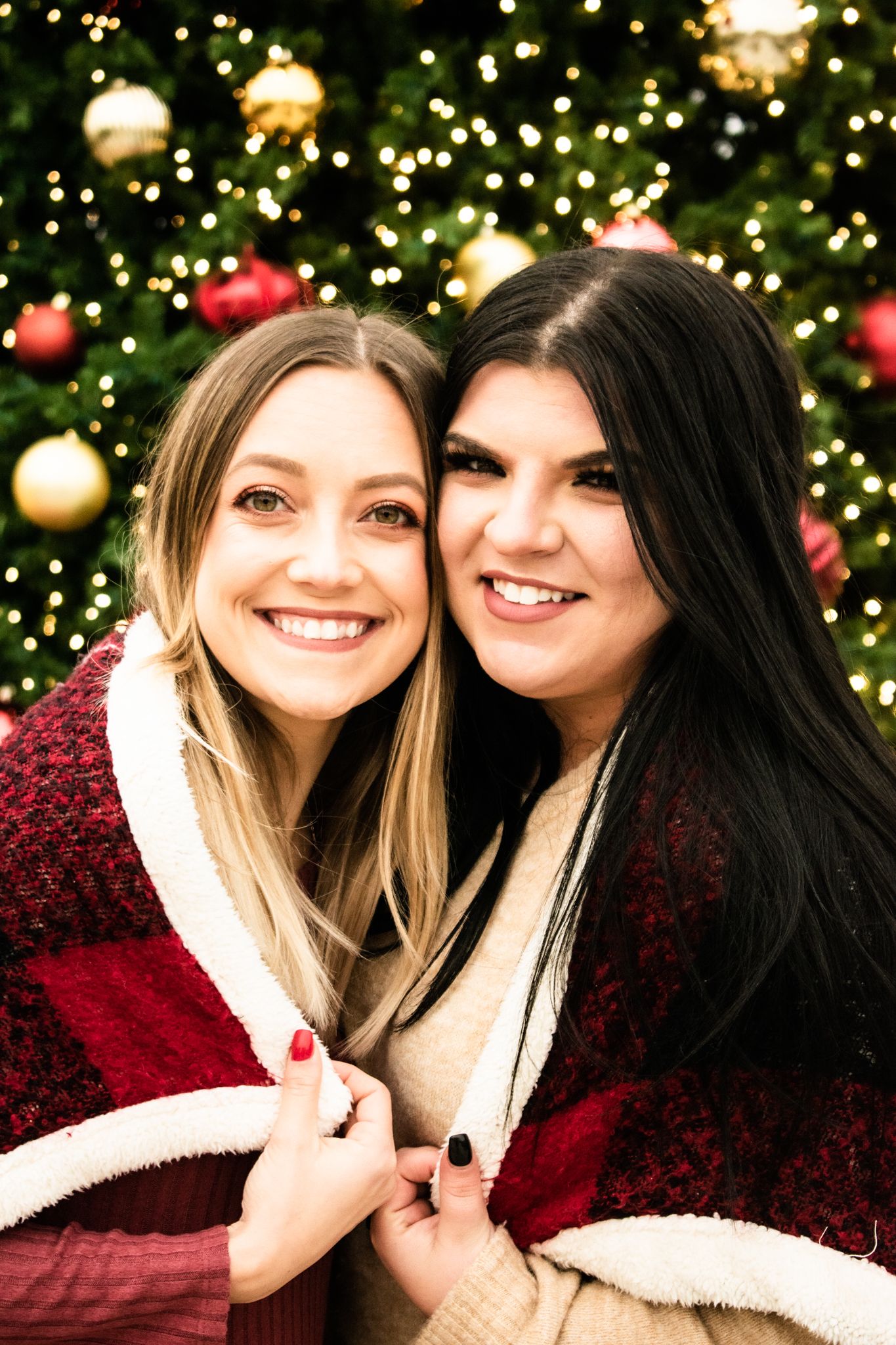 Two friends wrapped in plaid blankets smile together in front of holiday decorations.