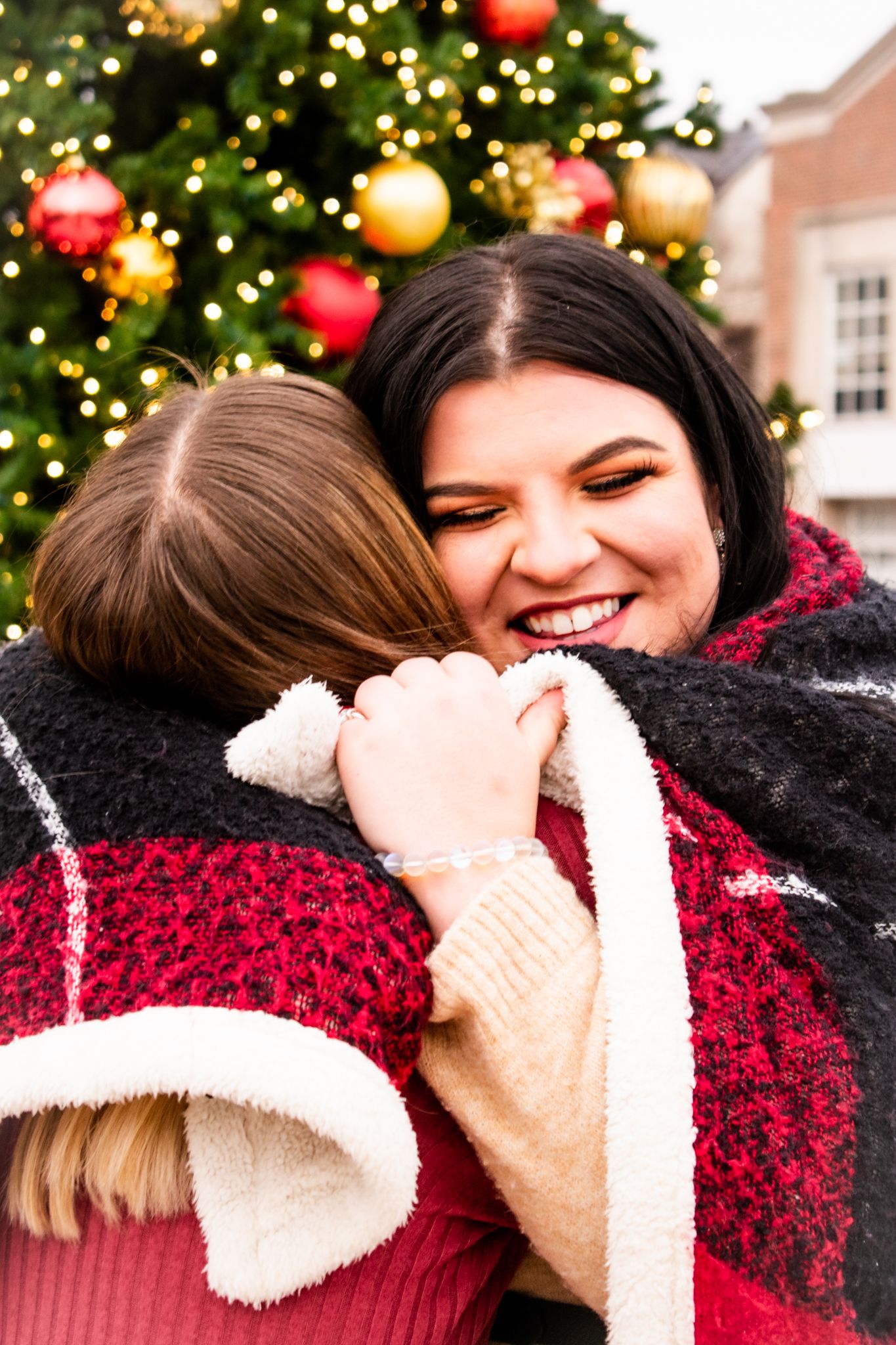 Friends embrace warmly in front of a festive Christmas tree with red and gold ornaments.