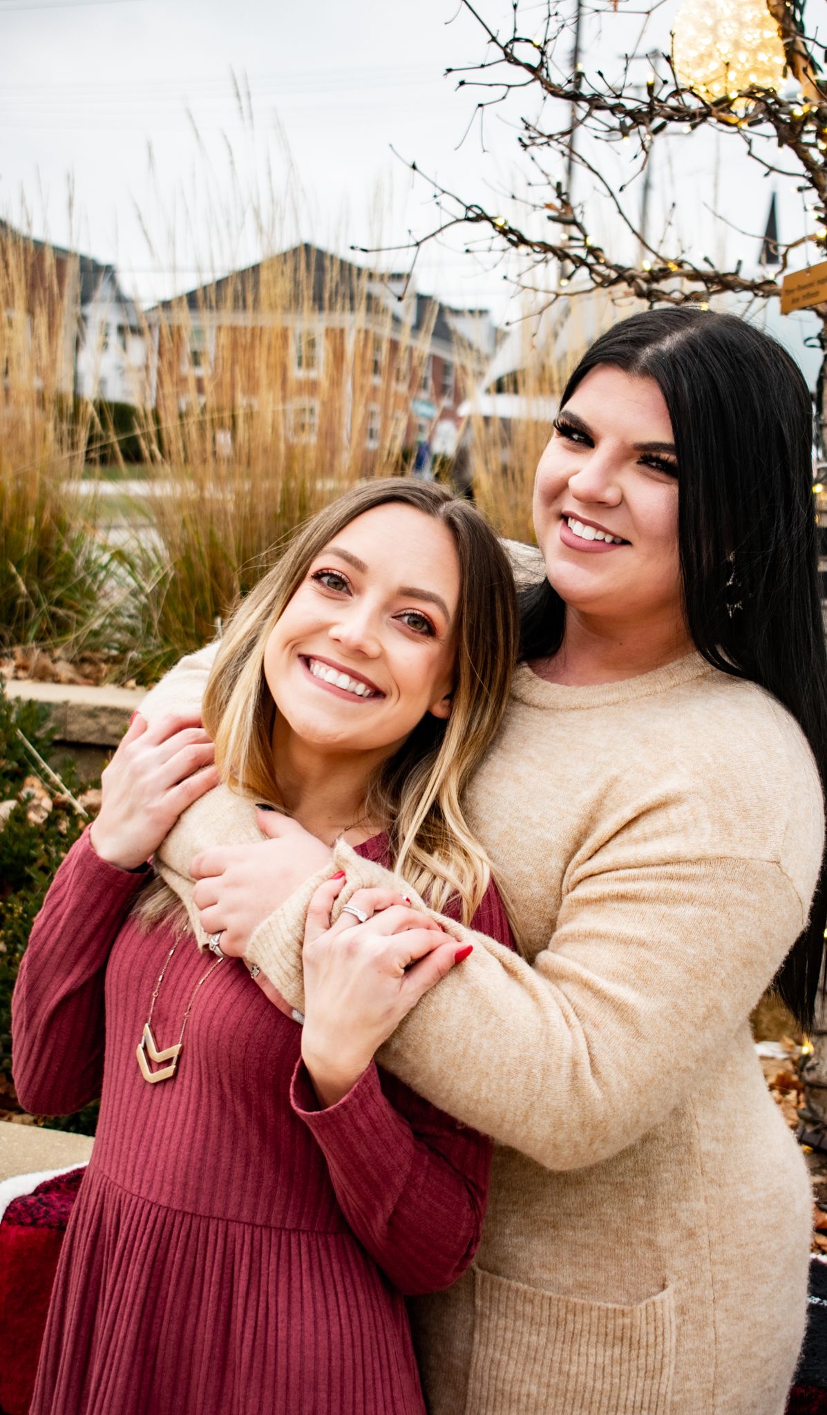 Friends embrace while posing outdoors in burgundy and beige winter outfits.