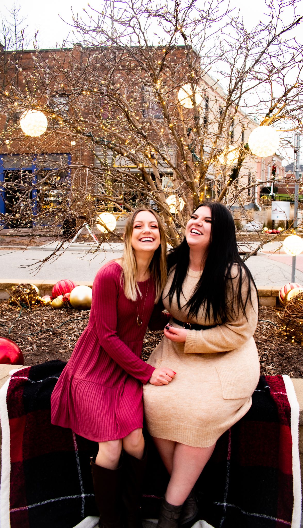 Two friends sitting on a blanket outdoors sharing laughs under twinkling holiday lights.