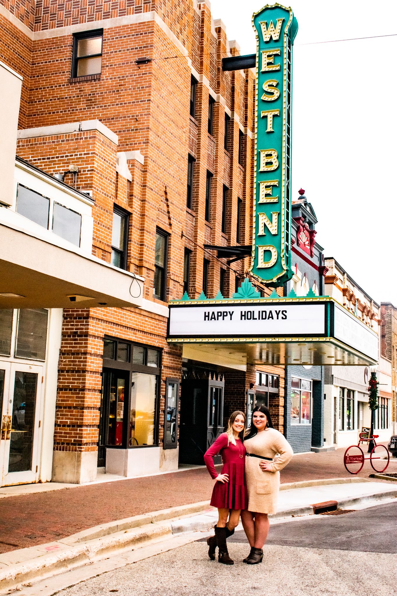 Two friends posing in front of historic West Bend theater with holiday marquee signage.
