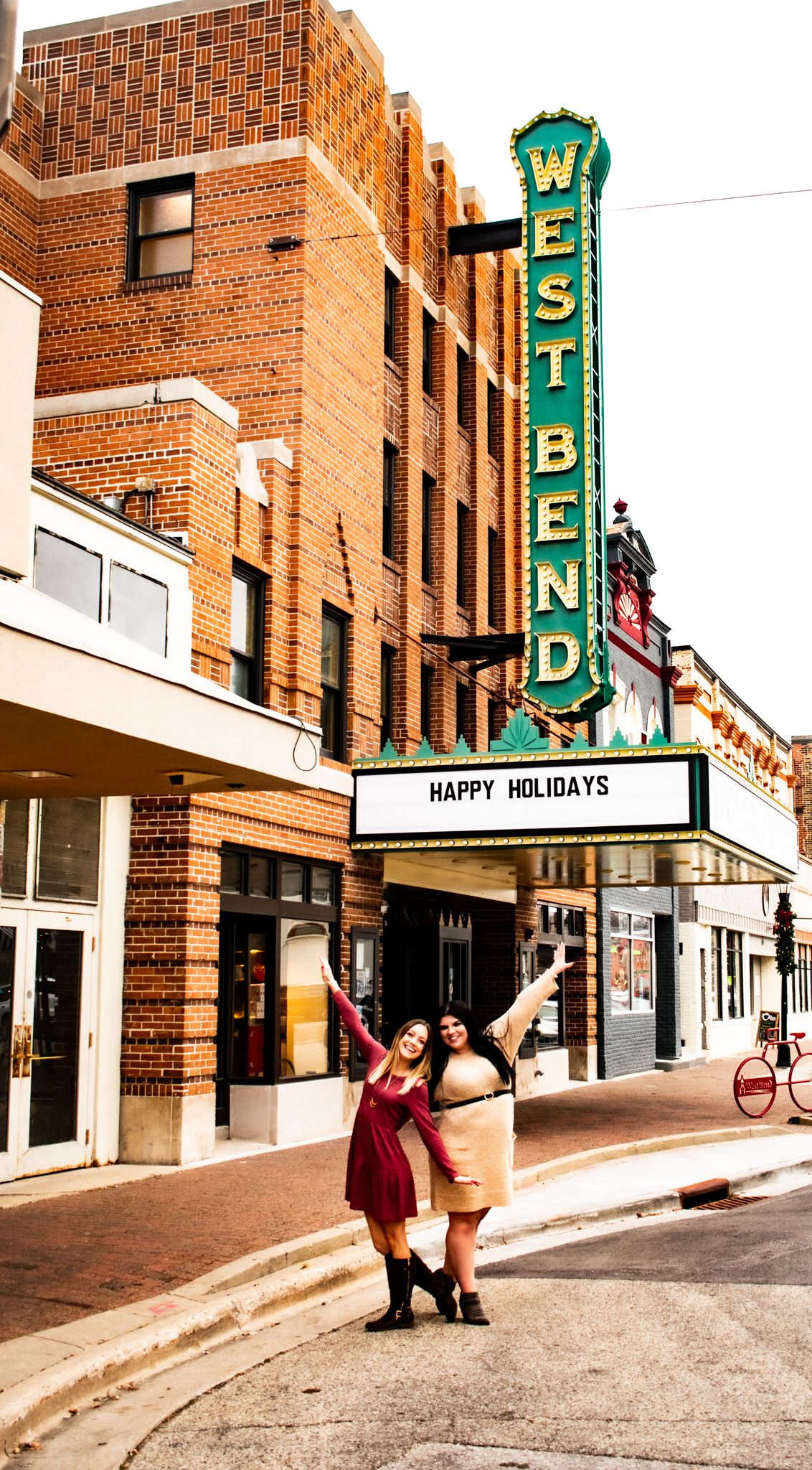 Two friends celebrating under the vintage West Bend theater marquee during holiday season.