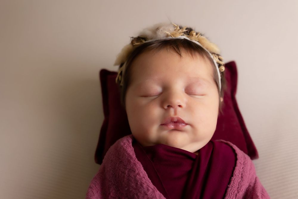 Sleeping baby wearing delicate headband wrapped in burgundy fabric against neutral backdrop.