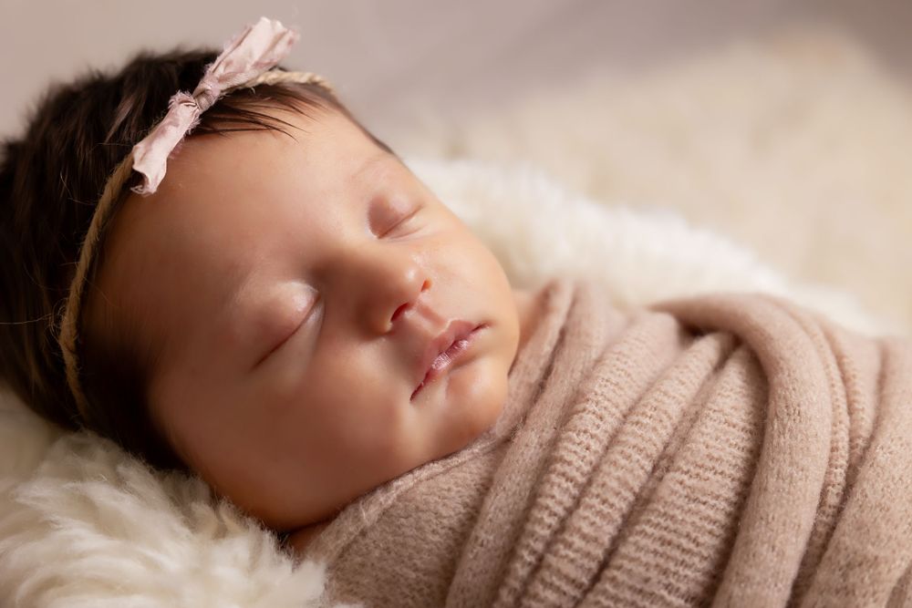 Serene newborn portrait with pink bow headband sleeping against cream fur backdrop.