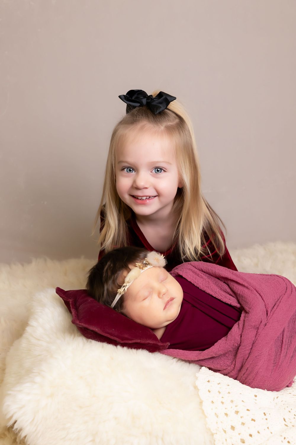 Young girl in red velvet dress with black bow poses with sleeping baby on white fur rug.