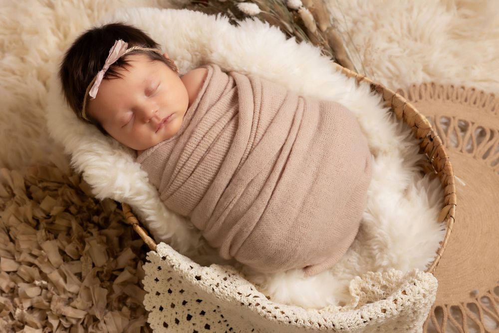 Swaddled sleeping infant in lace-trimmed basket on textured neutral background.