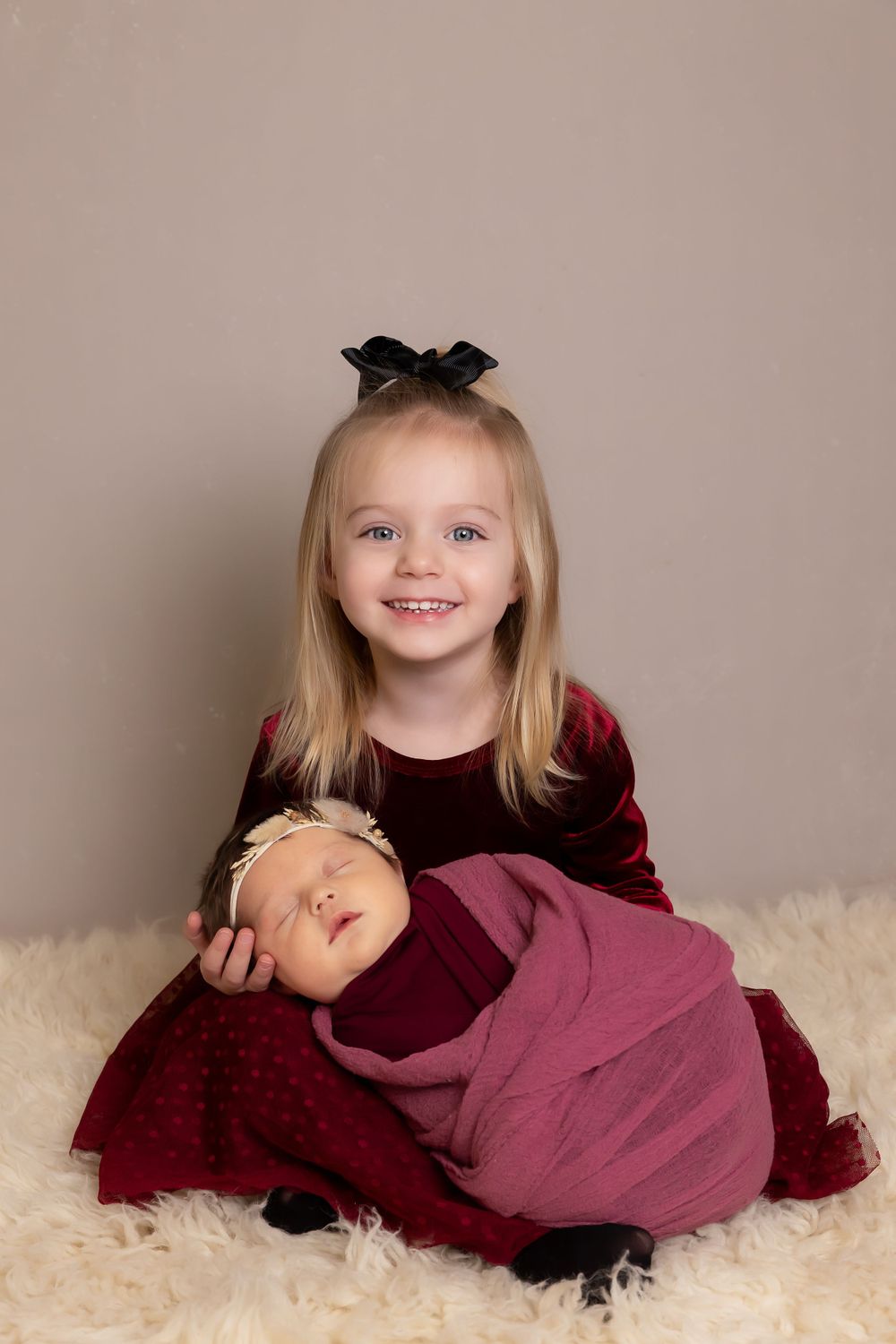 Girl with black bow and red dress poses with sleeping infant on plush white rug.
