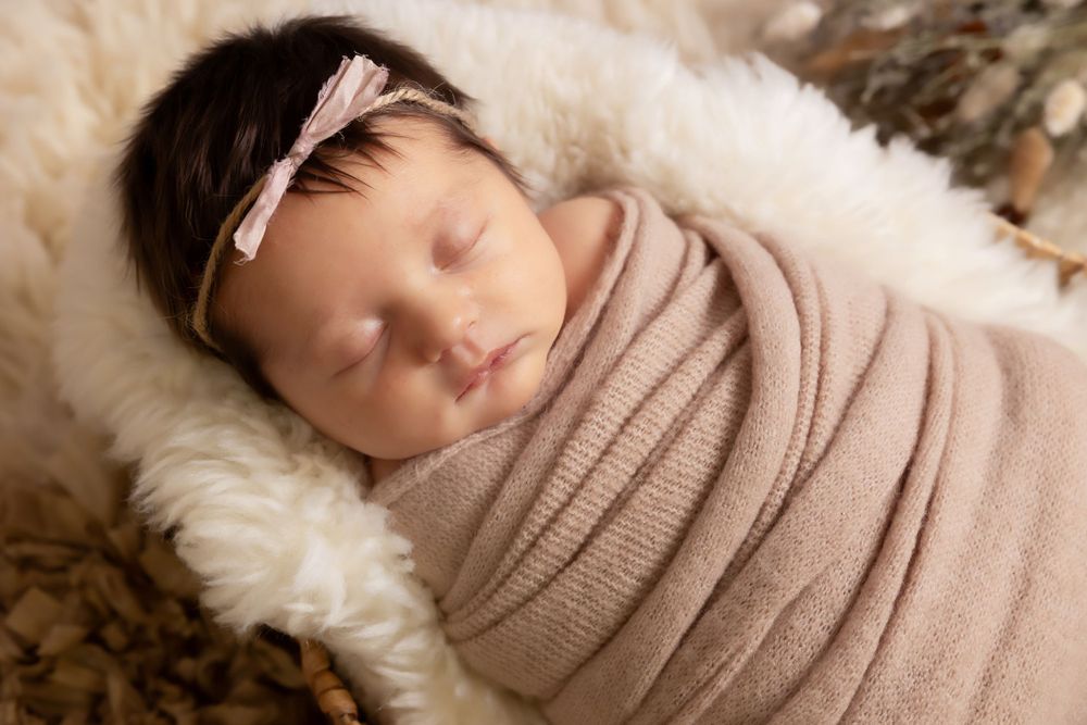 Sleeping newborn wrapped in beige knit swaddle resting on white fur with pink headband.