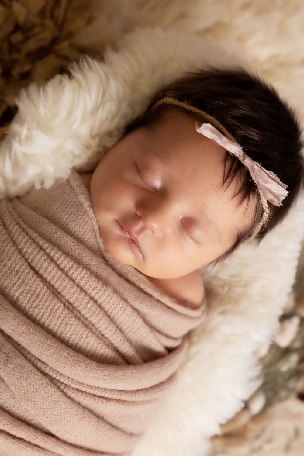 Close-up of peaceful newborn in tan wrap sleeping on soft white fur background.