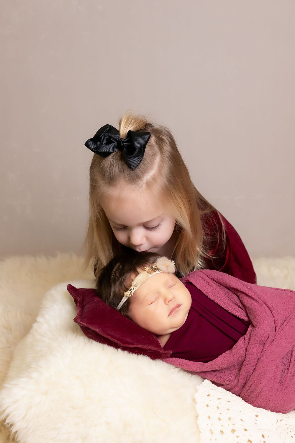Child in red dress leans over to kiss sleeping baby wrapped in burgundy blanket.