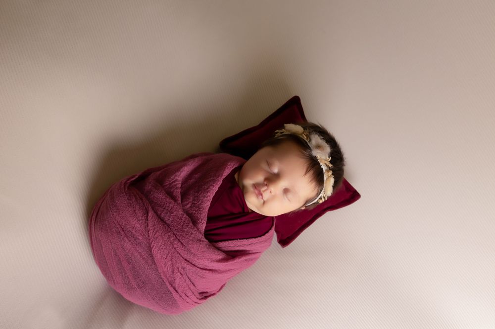 Newborn baby wearing headband sleeps peacefully wrapped in burgundy swaddle.