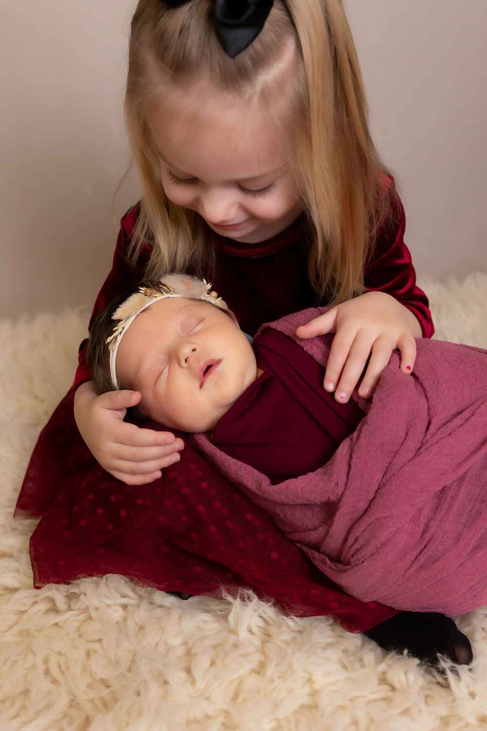 Girl in velvet dress tenderly holding sleeping baby wrapped in burgundy blanket.