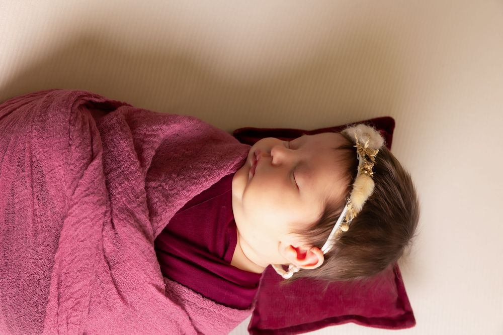 Close-up of sleeping baby with headband wrapped in burgundy blanket against neutral background.