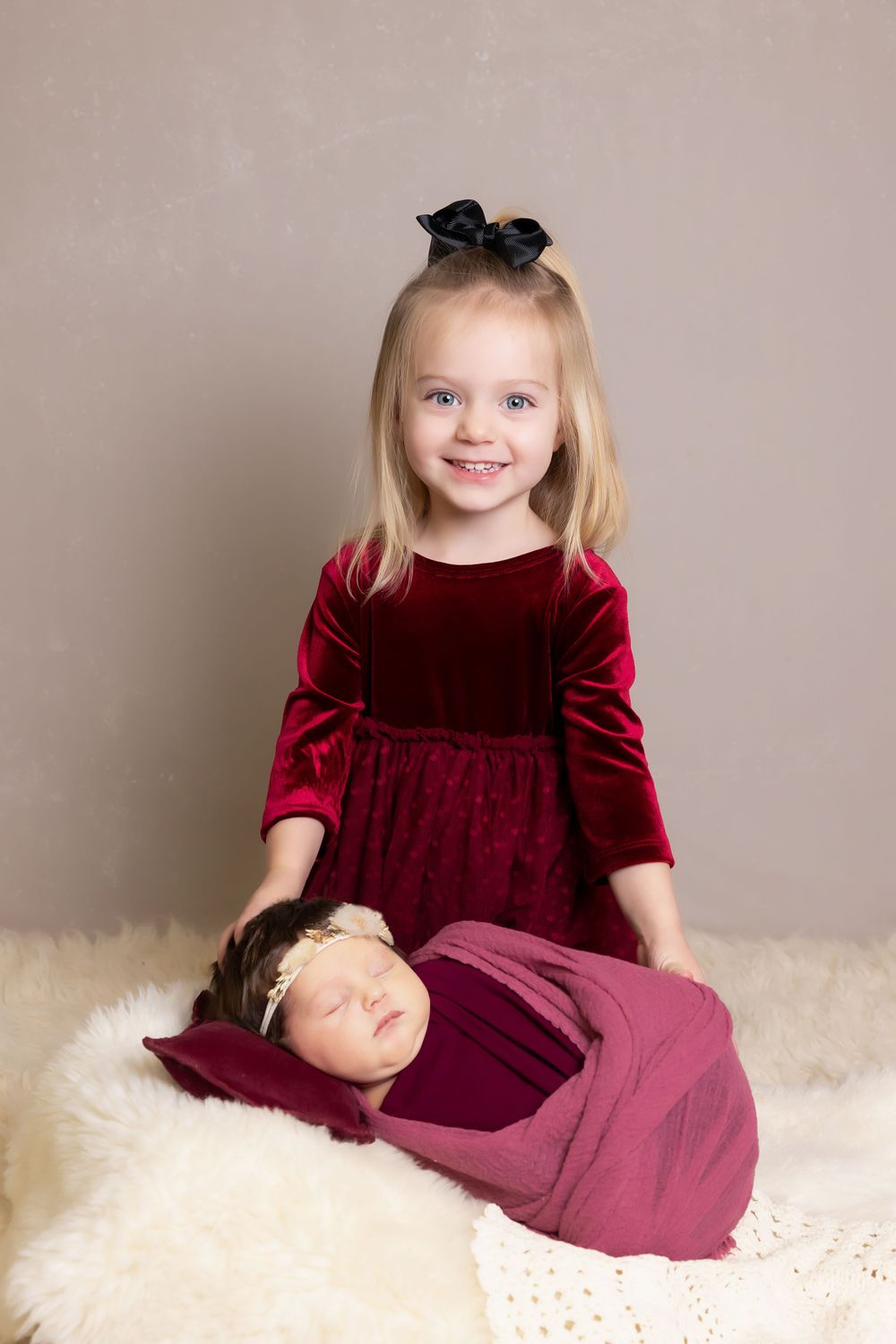 Girl in red velvet dress sitting with sleeping baby on fluffy white rug.